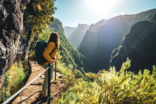 Backpacker woman enjoying scenic view from below large rock wall along water channel at steep cliff through Madeira's rainforest. Levada of Caldeirão Verde, Madeira Island, Portugal, Europe.