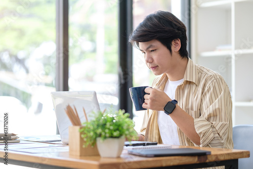 Image of young asian man freelancer drinking coffee and reading email on laptop.