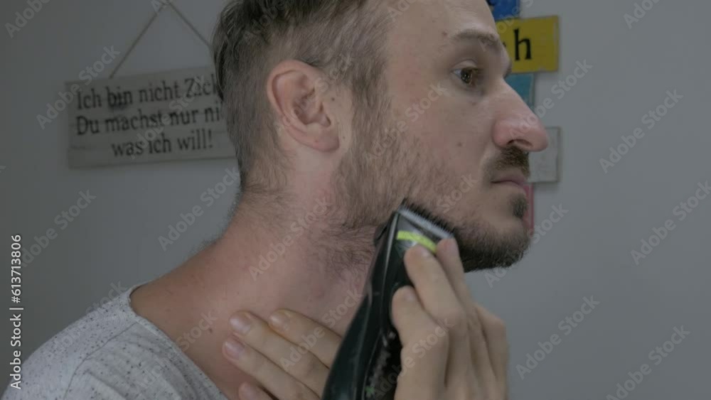 A Caucasian male is trimming his facial hair with an electric shaver to