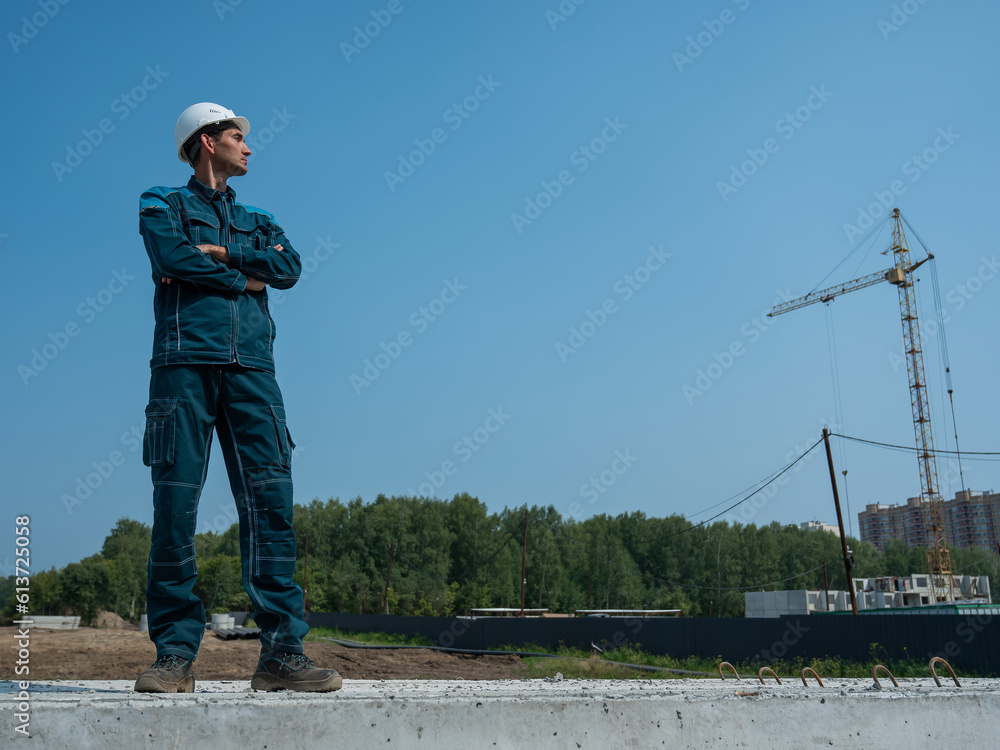 A builder in work clothes and a helmet stands on a construction site against the background of a construction crane. A builder in work clothes and a helmet stands on a construction site against the background of a construction crane.