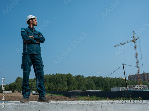A builder in work clothes and a helmet stands on a construction site against the background of a construction crane. 