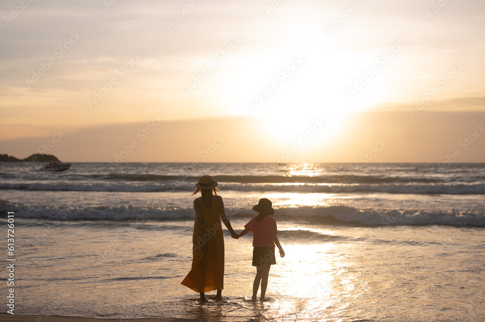 Young mother with her child walk on the beach of a beautiful coast at the silhouette sunset.