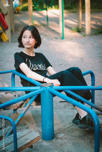 Portrait of a positive twelve-year-old teenage girl walking in the yard at the playground and riding a carousel. Summer day. Portrait of a child.