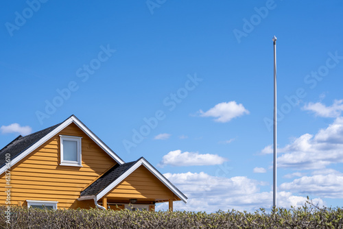 Norwegian landscape with a green hedge behind a yellow wooden building facade with a white window, next to the house is a white flagpole with beautiful clouds behind it