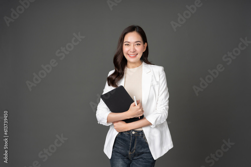 Asian female executive with long hair cute smile holding tablet and pen for work crossed arms, powerful posture Wearing a white suit, jeans and standing in a gray studio setting photo
