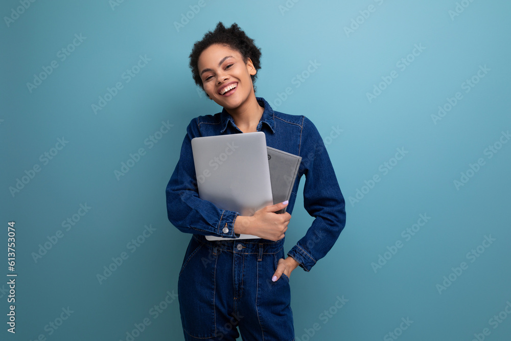 young happy brunette latin woman dressed in blue denim overalls studying remotely as a programmer and holding a laptop in her hands