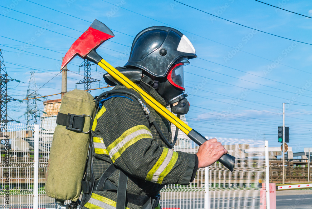 Naklejka premium A firefighter in a helmet and breathing apparatus put an assault axe on his shoulder. A firefighter in protective clothing and a mask on the side. A lifeguard with a fire axe. Rescue service.