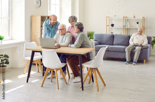 Senior people use computer in retirement home. Group of old men and women gather at table with laptop device, watch entertainment movie or make video call while other man reads book in background