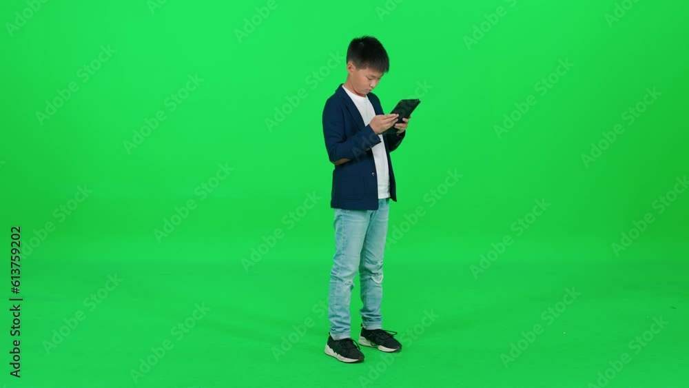 Asian schoolboy in a suit standing on a green background and using a screen tablet, reads an e-book, template on a chromakey. Asian schoolboy in a suit standing on a green background and using a screen tablet, reads an e-book, template on a chromakey.
