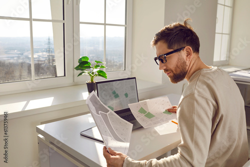 Portrait of serious focused attractive architect in process of work, sitting at desk at his workplace in office. Young engineer with glasses with cadastral maps and projects sitting in front of laptop photo