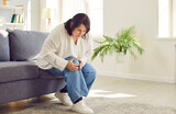 Portrait of young plus size overweight woman holding a knee suffering from osteoarthritis or arthritis sitting on a sofa in the living room at home. Obesity, chronic disease, health care concept.