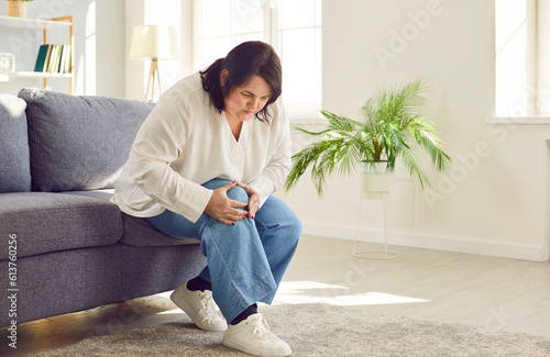 Portrait of young plus size overweight woman holding a knee suffering from osteoarthritis or arthritis sitting on a sofa in the living room at home. Obesity, chronic disease, health care concept.