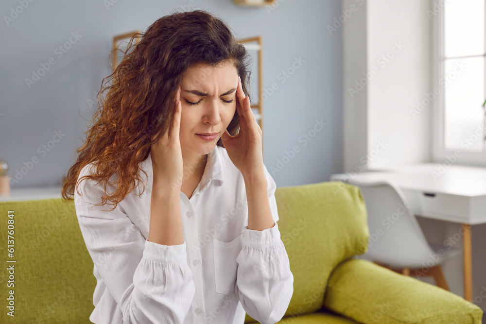 Depressed young woman with headache sitting on sofa. Attractive woman suffering from vertigo, dizziness, migraine, stress, overwork. Sick girl massaging temples with her hands Depressed young woman with headache sitting on sofa. Attractive woman suffering from vertigo, dizziness, migraine, stress, overwork. Sick girl massaging temples with her hands