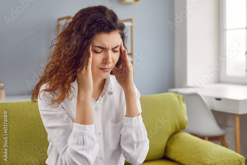 Depressed young woman with headache sitting on sofa. Attractive woman suffering from vertigo, dizziness, migraine, stress, overwork. Sick girl massaging temples with her hands