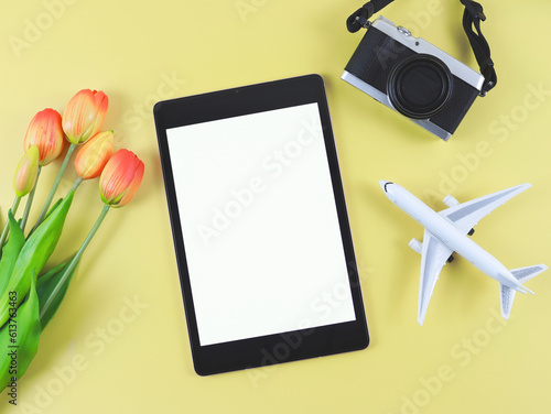 flat lay of digital tablet with blank white screen, tulip flowers, airplane model and digital camera isolated on yellow background.