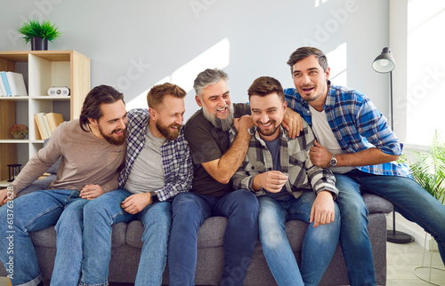 Portrait of a male bearded friends sitting on sofa, smiling and having fun at home in the living room. Photo of group of brutal friendly men in casual clothes hugging indoors on the party.