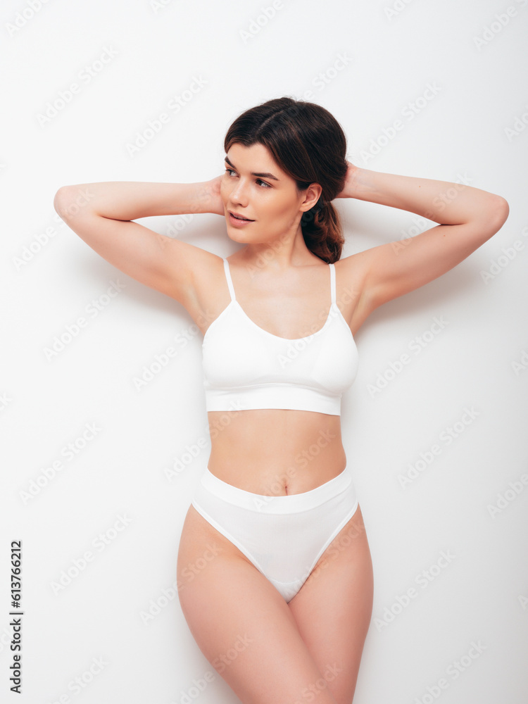 Portrait of young beautiful sexy woman in white lingerie. Smiling carefree model wearing pure underwear. Hot brunette posing near wall in studio interior. Perfect body. Slim and fit Portrait of young beautiful sexy woman in white lingerie. Smiling carefree model wearing pure underwear. Hot brunette posing near wall in studio interior. Perfect body. Slim and fit