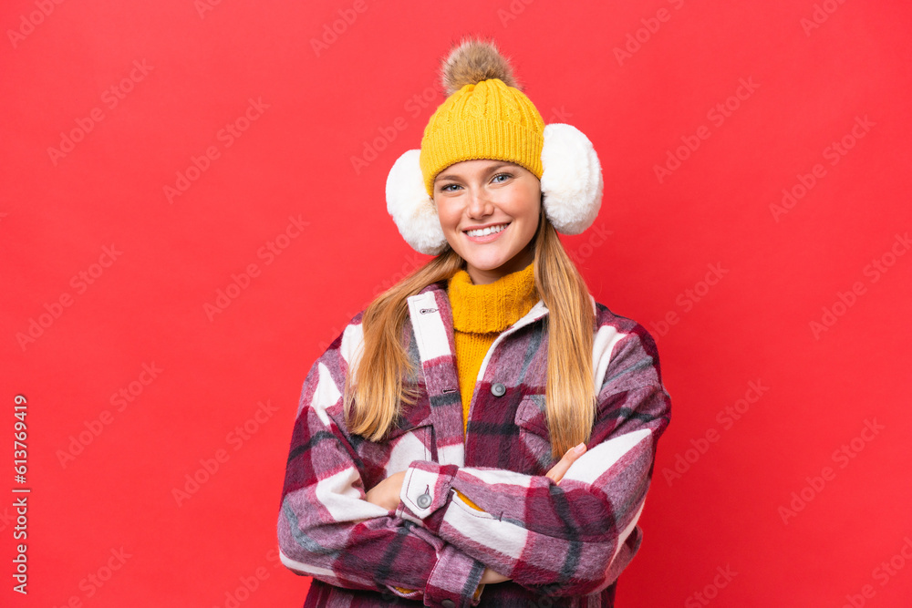 Young beautiful woman wearing winter muffs isolated on red background keeping the arms crossed in frontal position Young beautiful woman wearing winter muffs isolated on red background keeping the arms crossed in frontal position