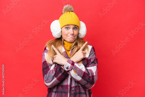 Young beautiful woman wearing winter muffs isolated on red background pointing to the laterals having doubts