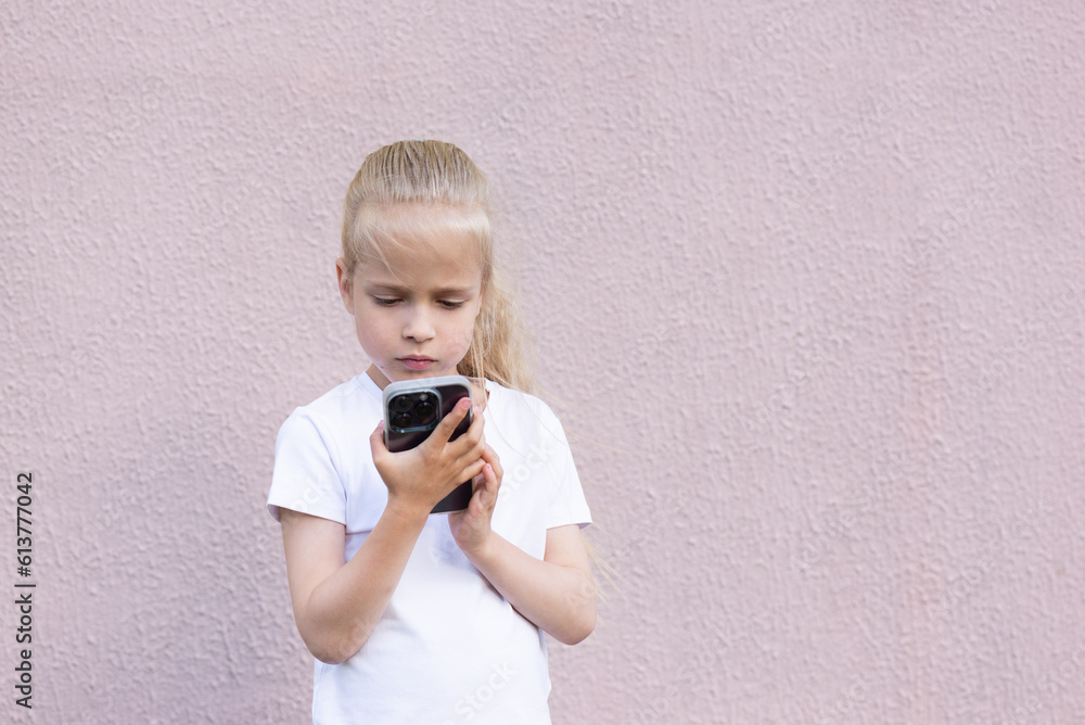 Little blonde girl looks into a smartphone against a pink wall. Little blonde girl looks into a smartphone against a pink wall.