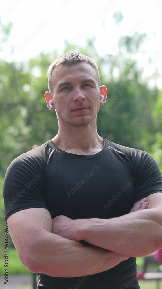 Slow motion low angle of confident male athlete wearing black sportswear and earbuds while standing on sports ground crossing arms and looking at camera Vertical video Slow motion low angle of confident male athlete wearing black sportswear and earbuds while standing on sports ground crossing arms and looking at camera Vertical video