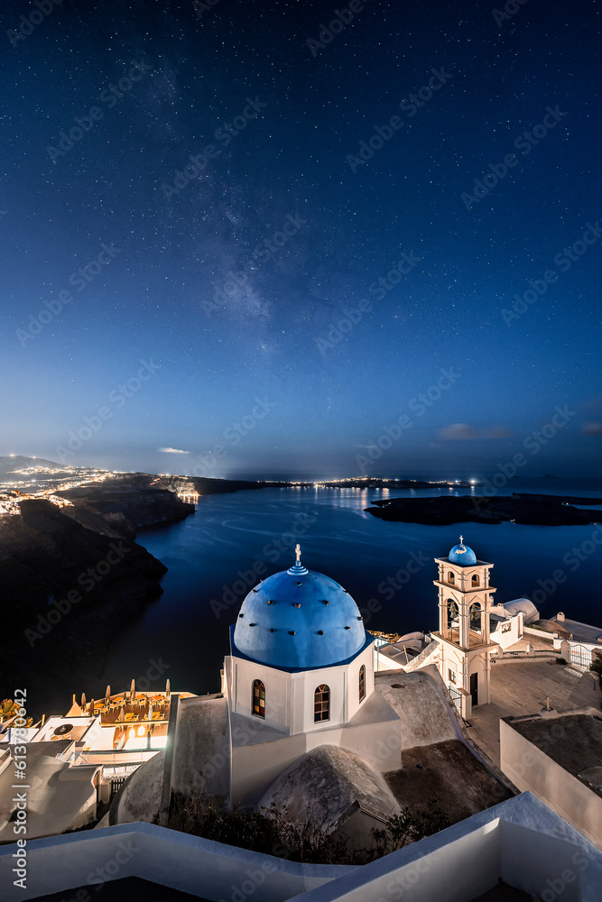 Fototapeta premium Night time image with stars and milky way of white and blue church with view on the caldera cliff and cruise ships on the Greek island of Santorini