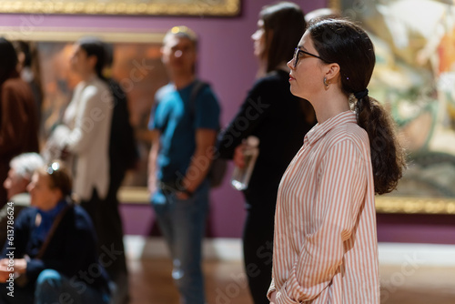 Side view of young Caucasian woman wearing glasses and looking at exhibition. People and masterpieces in background. Concept of Museum Day