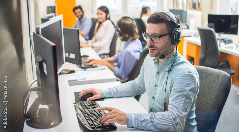 Male customer service representative with headset working in call center office with colleagues in row. - obrazy, fototapety, plakaty 