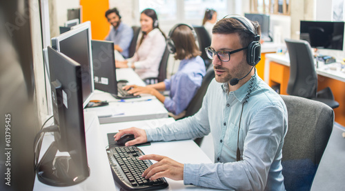 Male customer service representative with headset working in call center office with colleagues in row.