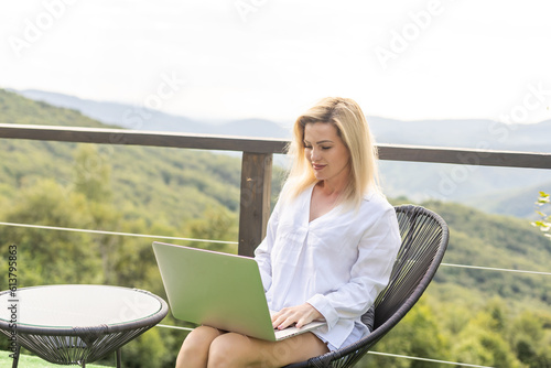 woman sitting on hotel balcony with laptop computer while working remote on freelance. mountains view