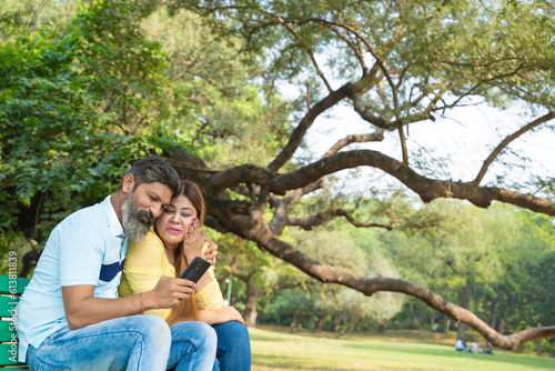 Happy indian couple sitting at garden and watching some in smartphone. © Niks Ads