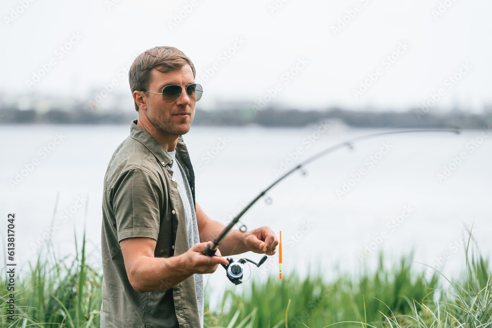 Standing near the lake. Handsome man in sunglasses is fishing outdoors Standing near the lake. Handsome man in sunglasses is fishing outdoors