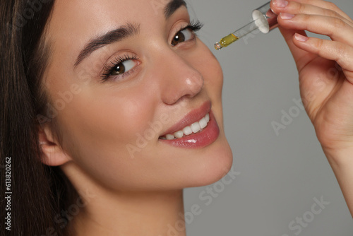 Beautiful young woman applying essential oil onto face on grey background, closeup