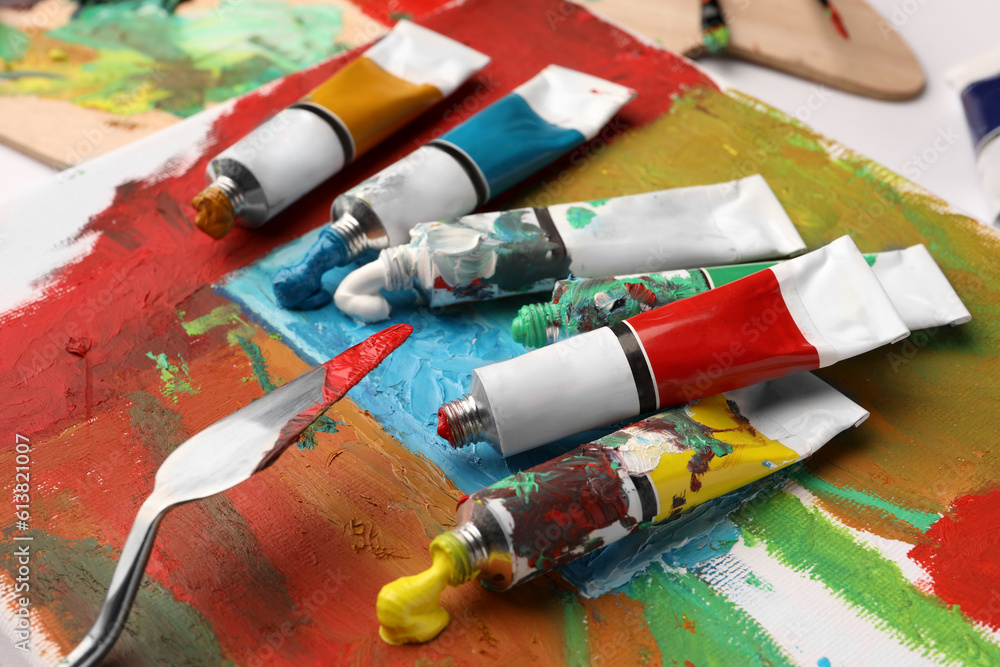 Tubes of colorful oil paints, spatula and canvas with abstract painting on table, closeup