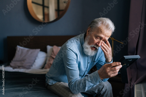 Side view of sad bearded gray-haired mature adult male looking at picture of wife in frame and crying, sitting on couch in dark room. Concept of nostalgia, grief, longing, loneliness in old age.