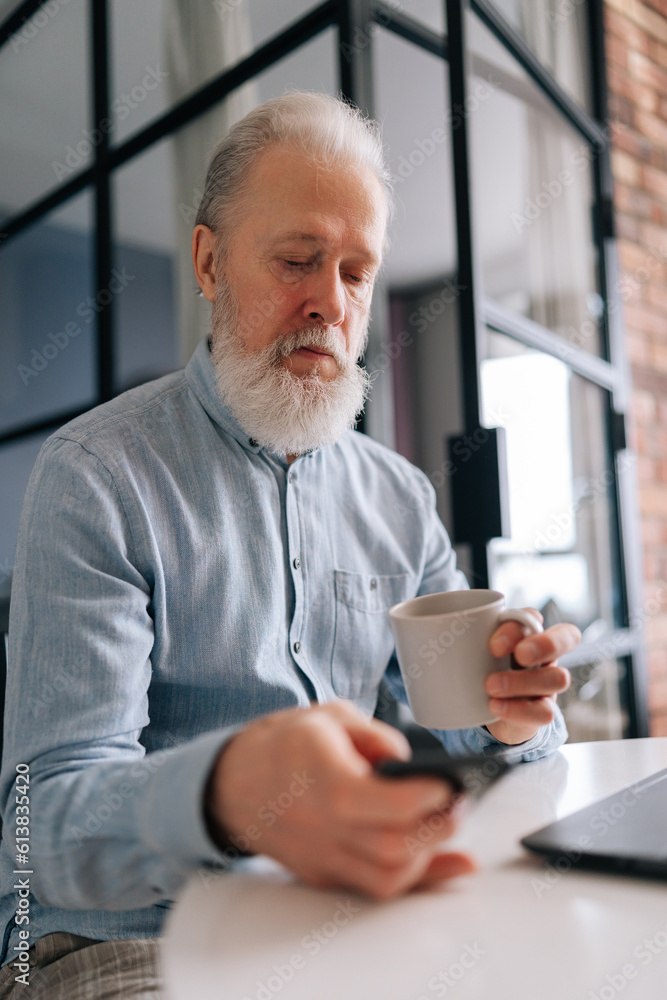 Vertical portrait of elderly mature male with grey hair working on laptop computer and using smartphone, holding cup of coffee, sitting at table in kitchen room in morning, looking device screen. Vertical portrait of elderly mature male with grey hair working on laptop computer and using smartphone, holding cup of coffee, sitting at table in kitchen room in morning, looking device screen.