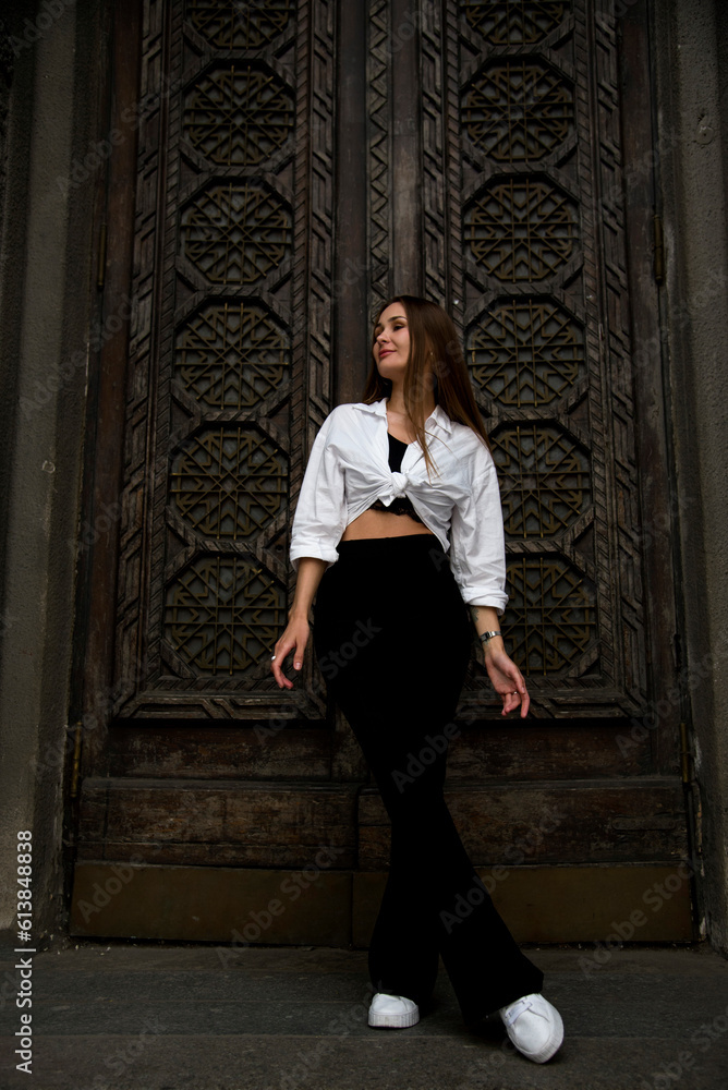 portrait of a beautiful brunette woman with long hair staying near wooden carved door in white shirt and black trousers 