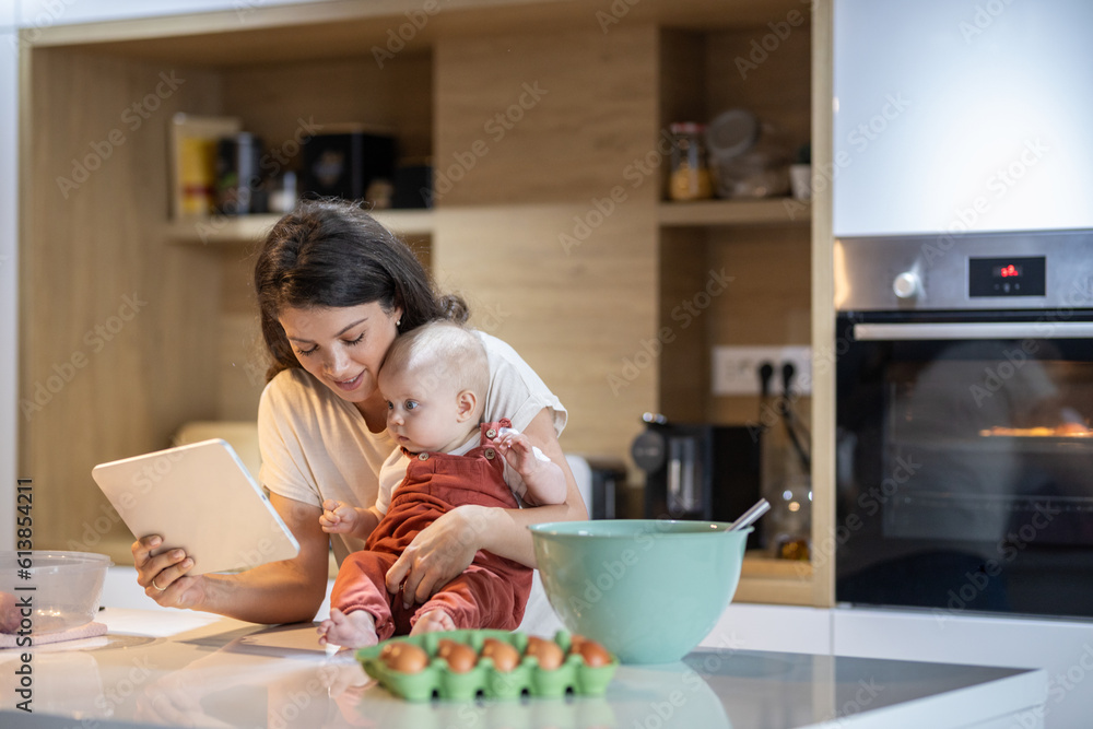 Young mom shows her baby something on her tablet as she holds her on the kitchen counter