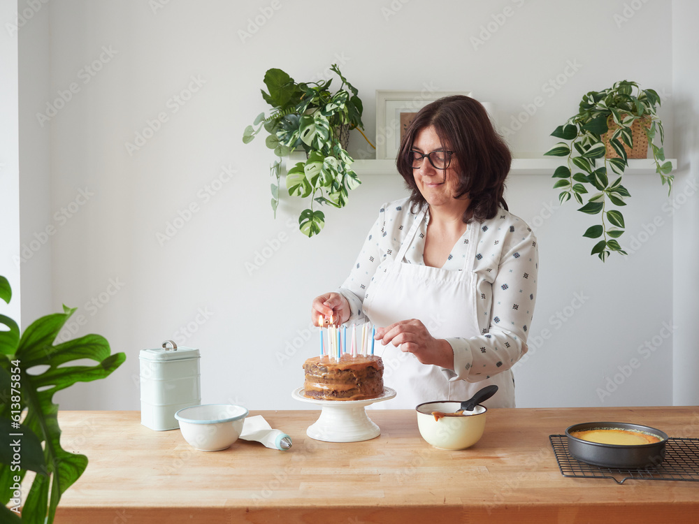 smiling woman lighting candles on chocolate layer cake on wooden countertop with other decorating utensils smiling woman lighting candles on chocolate layer cake on wooden countertop with other decorating utensils