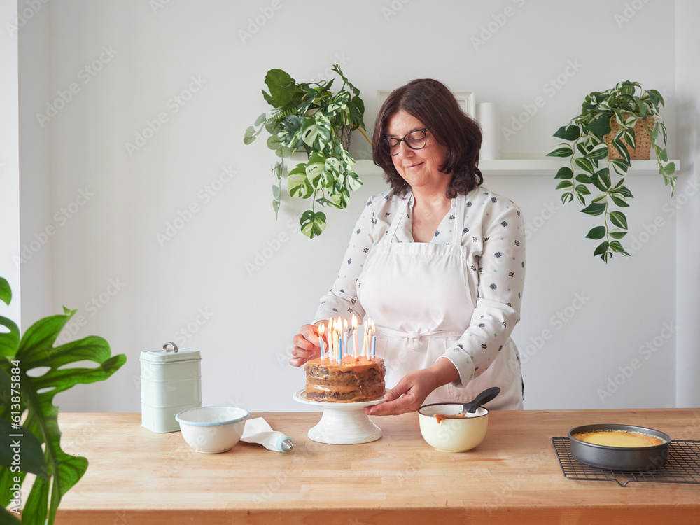 smiling woman placing lighted candles on chocolate cream birthday cake at home on wooden countertop with other decorating utensils smiling woman placing lighted candles on chocolate cream birthday cake at home on wooden countertop with other decorating utensils