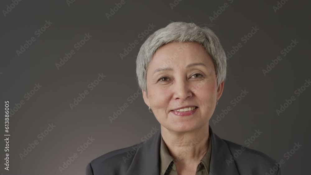 Chest up portrait of confident mature Asian woman with short grey hair taking off eyeglasses and smiling at camera standing on graphite studio background Chest up portrait of confident mature Asian woman with short grey hair taking off eyeglasses and smiling at camera standing on graphite studio background