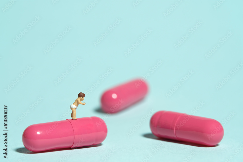 Miniature tiny people toy figure photography. A toddler baby walking above pink medicine pill tablet capsule. Isolated on blue background Miniature tiny people toy figure photography. A toddler baby walking above pink medicine pill tablet capsule. Isolated on blue background