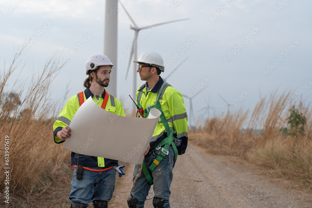 Two male windmill engineer in uniform and helmet safety discussing inspection and maintenance of wind turbine in wind farms to generate electrical energy, Renewable power energy Two male windmill engineer in uniform and helmet safety discussing inspection and maintenance of wind turbine in wind farms to generate electrical energy, Renewable power energy