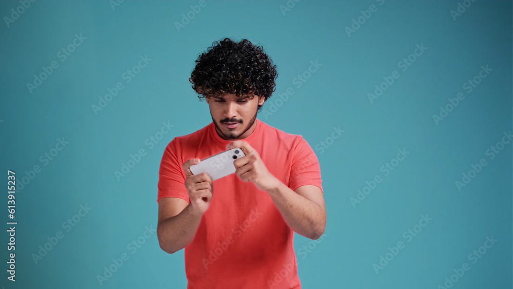 Excited happy young swarthy man gamer player using smartphone, playing game in mobile videogame app or watching digital video stream holding cell phone isolated on blue studio background. Excited happy young swarthy man gamer player using smartphone, playing game in mobile videogame app or watching digital video stream holding cell phone isolated on blue studio background.