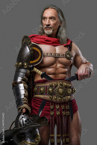 Distinguished gladiator with a stylish silver beard and luscious locks wears lightweight armor, holding gladiator helmet with feathered plume as he stands confidently against grey backdrop