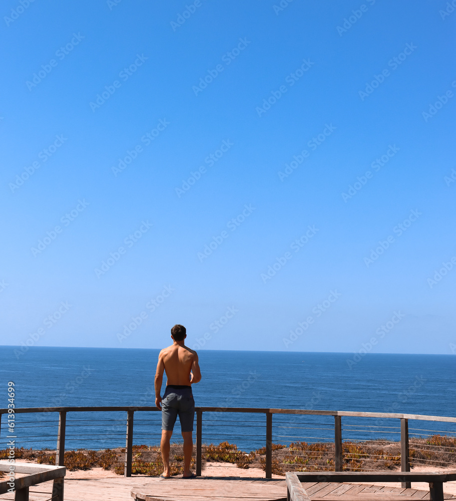 Young man traveler contemplating in a peaceful place in front of the ocean viewed from the back. Portugal Solo traveler.