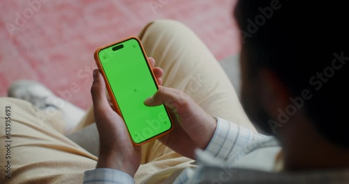 A man in casual clothes scroll the green screen of a mobile phone while sitting on the couch at home. Top view, no face, close-up of a man's hands