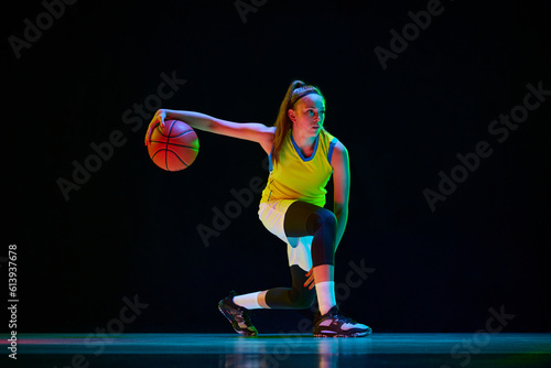 Young girl, professional basketball player in motion, training, playing against black studio background in neon light. Concept of professional sport, action and motion, game, competition, hobby, ad