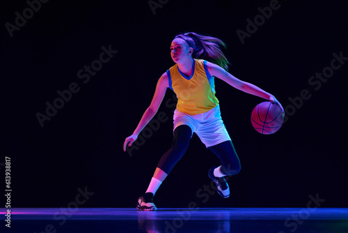 Dynamic image of young girl, basketball player in motion with ball, playing over black studio background in neon light. Concept of professional sport, action and motion, game, competition, hobby, ad
