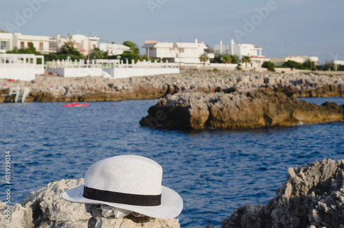 White hat on the rocks near the sea. In the blurry background a sea village (Santa Caterina, Salento, Italy)